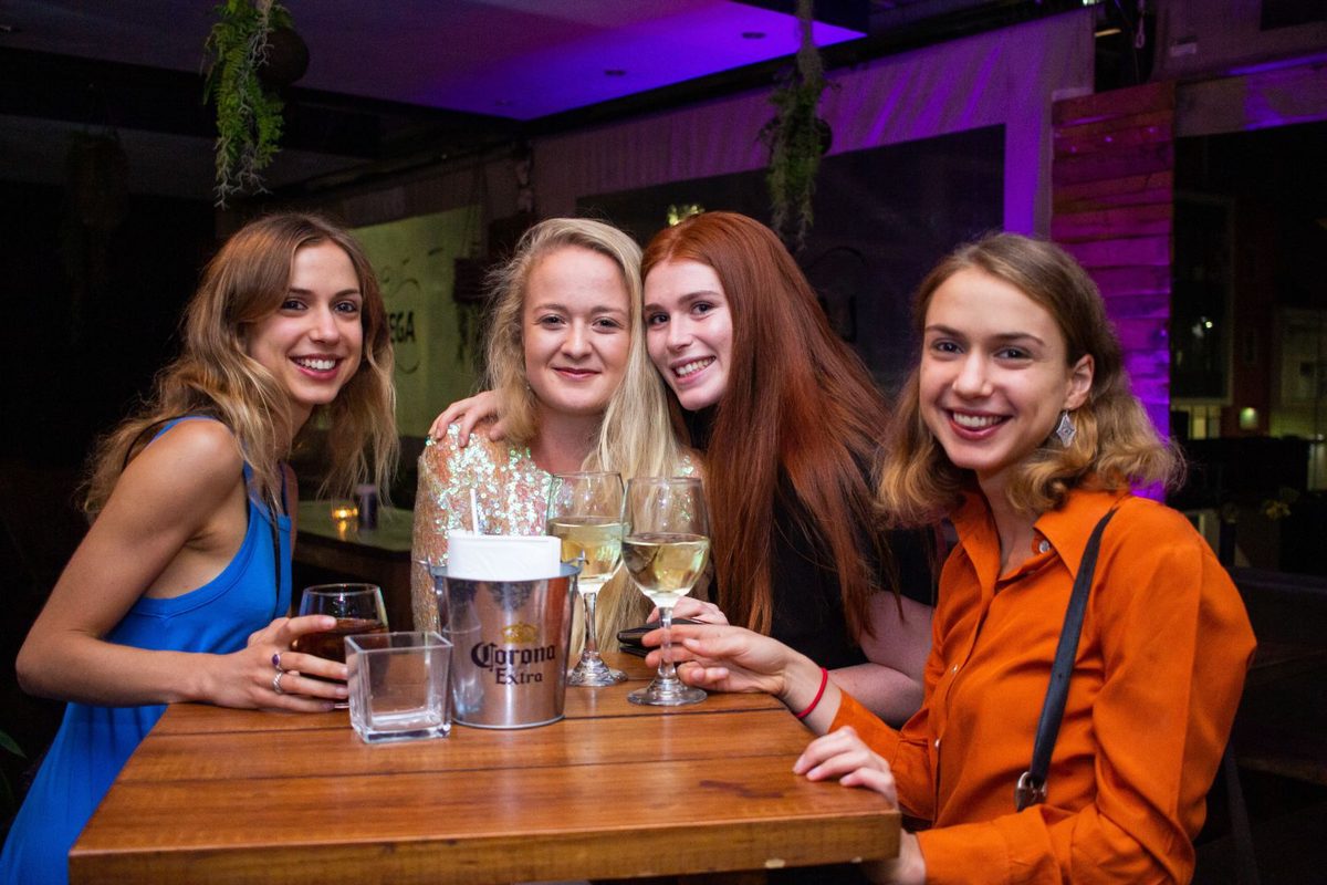 Four women smiling at camera, two holding wine glasses, one holding a Corona Extra bucket